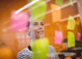 Mulher olhando para post-its rosas, amarelos e verdes em uma parede de vidro em escritório