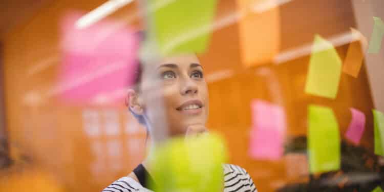 Mulher olhando para post-its rosas, amarelos e verdes em uma parede de vidro em escritório