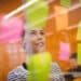 Mulher olhando para post-its rosas, amarelos e verdes em uma parede de vidro em escritório