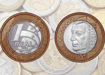 Frente e verso da moeda de 1 Real comemorativa do Centenário de Juscelino Kubitschek (2002) sobre um fundo de várias moedas desfocadas.