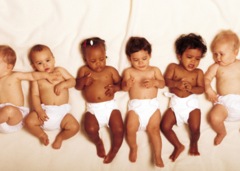 Seis bebês de várias etnias deitados lado a lado usando fraldas brancas em fundo claro.