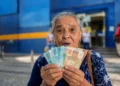 Idosa brasileira segura cédulas de real em frente a uma agência bancária, representando o pagamento dos benefícios do INSS de agosto para segurados que recebem acima de um salário mínimo.