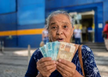 Idosa brasileira segura cédulas de real em frente a uma agência bancária, representando o pagamento dos benefícios do INSS de agosto para segurados que recebem acima de um salário mínimo.