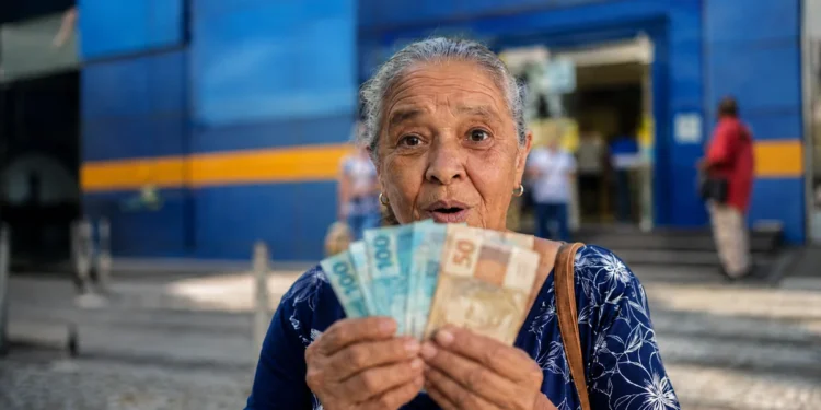 Idosa brasileira segura cédulas de real em frente a uma agência bancária, representando o pagamento dos benefícios do INSS de agosto para segurados que recebem acima de um salário mínimo.
