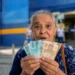 Idosa brasileira segura cédulas de real em frente a uma agência bancária, representando o pagamento dos benefícios do INSS de agosto para segurados que recebem acima de um salário mínimo.