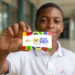 Estudante segurando cartão do programa Pé-de-Meia em frente à escola representando os pagamentos liberados pelo MEC.