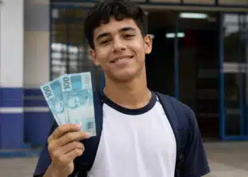 Estudante segura cédulas de R$ 100 em frente à escola, representando o pagamento do Pé-de-Meia para nascidos em novembro e dezembro.