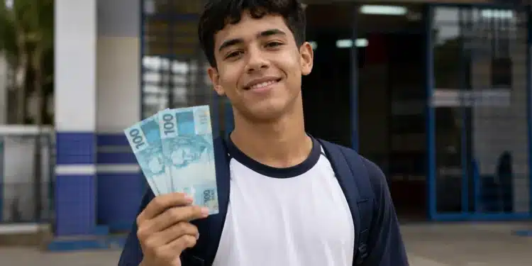 Estudante segura cédulas de R$ 100 em frente à escola, representando o pagamento do Pé-de-Meia para nascidos em novembro e dezembro.