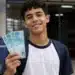 Estudante segura cédulas de R$ 100 em frente à escola, representando o pagamento do Pé-de-Meia para nascidos em novembro e dezembro.