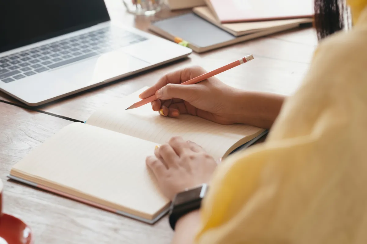 Concursos Mãos segurando lápis e escrevendo em caderno aberto sobre mesa de madeira com notebook ao fundo