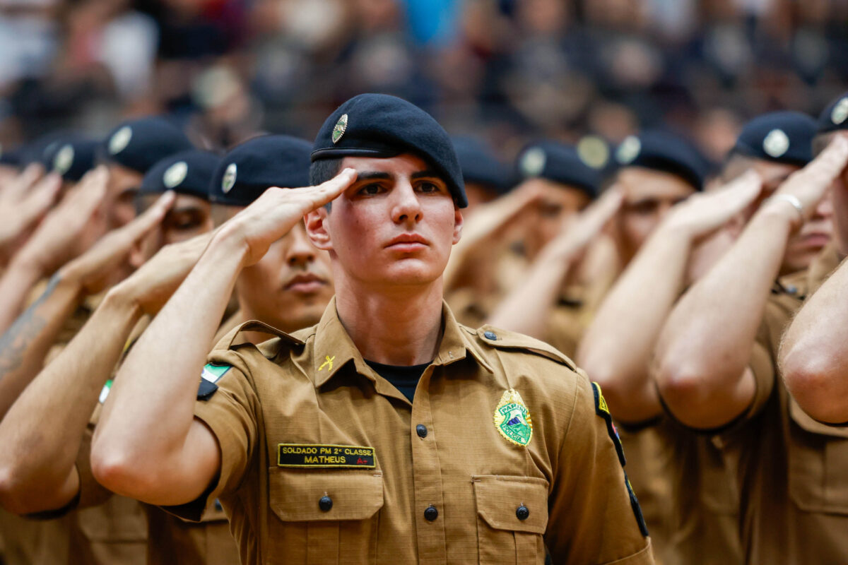 Policiais militares do Paraná em uniforme marrom e boina preta fazendo saudação durante cerimônia oficial
