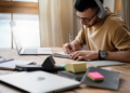 Homem com headset e óculos escrevendo em caderno ao lado de laptop aberto em mesa de madeira