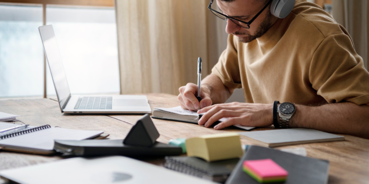 Homem com headset e óculos escrevendo em caderno ao lado de laptop aberto em mesa de madeira