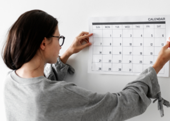 Mulher de cabelos escuros e óculos ajustando calendário branco com dias do mês na parede branca