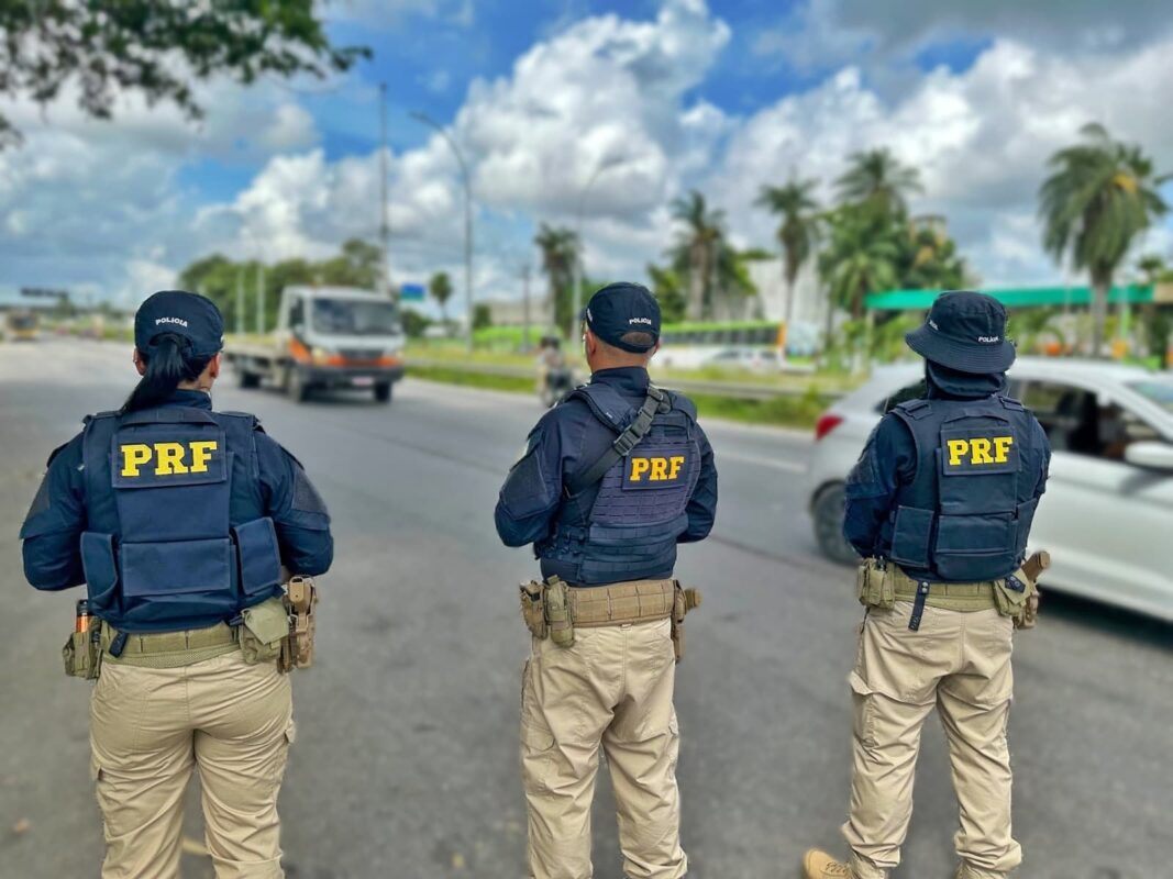 Concurso PRF Três policiais da PRF em operação, fardados com coletes à prova de balas, em rodovia sob céu azul com nuvens