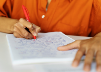 Mão escrevendo e corrigindo texto em caderno de folhas quadriculadas com caneta vermelha