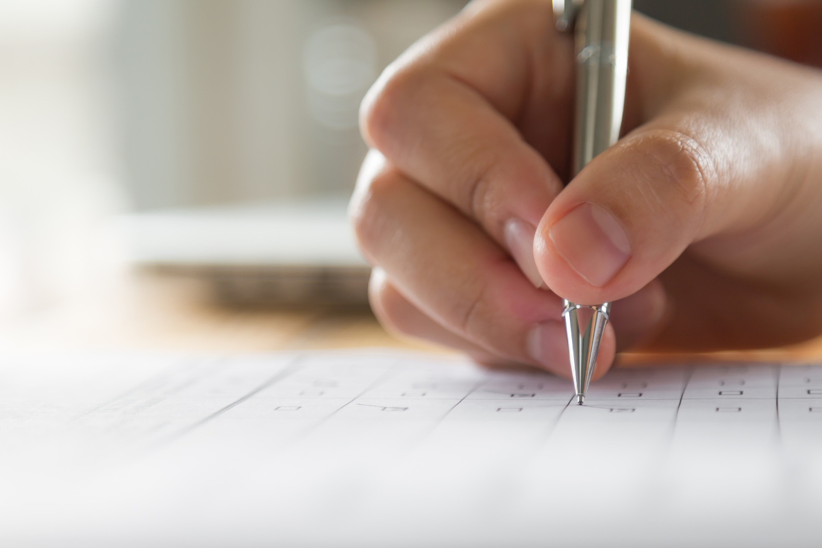 Mão segurando caneta prata preenchendo formulário com caixas para marcar em papel branco desfocado