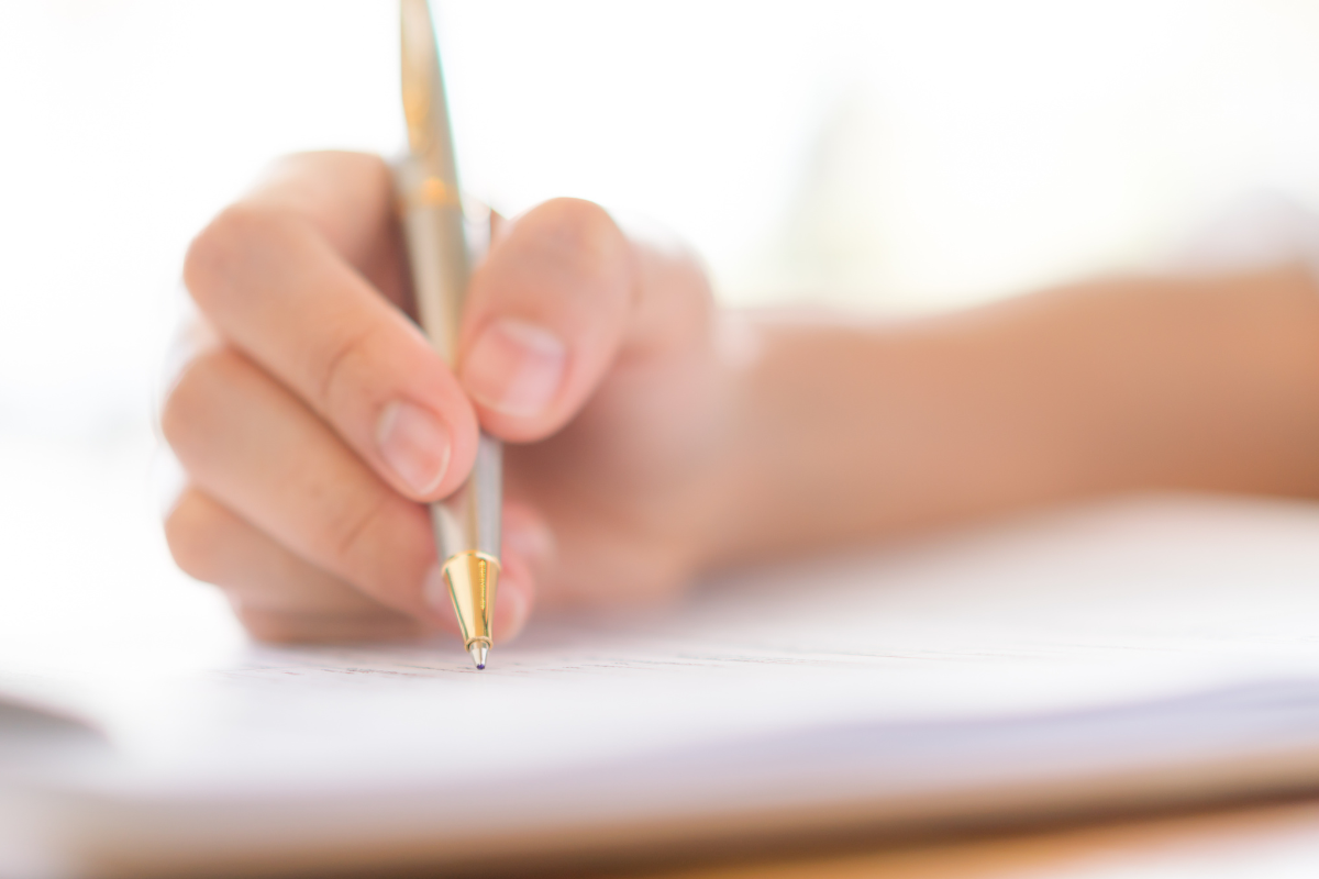 Mão segurando uma caneta dourada escrevendo em uma folha de papel branca com fundo desfocado