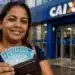 Mulher sorridente segurando carteira com notas de R$ 100 em frente a uma agência da Caixa, representando o Saque Calamidade do FGTS.