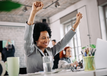 Mulher negra com cabelo afro e terno cinza levantando os braços em comemoração no ambiente de trabalho