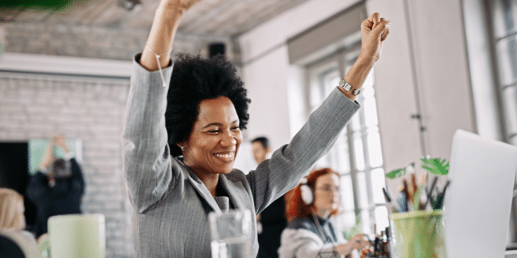 Mulher negra com cabelo afro e terno cinza levantando os braços em comemoração no ambiente de trabalho