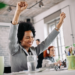 Mulher negra com cabelo afro e terno cinza levantando os braços em comemoração no ambiente de trabalho