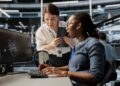 Duas especialistas em tecnologia, uma mulher negra sentada operando um computador e uma mulher branca em pé com um tablet, analisando juntas gráficos de dados complexos em um escritório moderno.