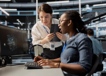 Duas especialistas em tecnologia, uma mulher negra sentada operando um computador e uma mulher branca em pé com um tablet, analisando juntas gráficos de dados complexos em um escritório moderno.