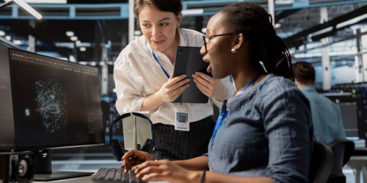 Duas especialistas em tecnologia, uma mulher negra sentada operando um computador e uma mulher branca em pé com um tablet, analisando juntas gráficos de dados complexos em um escritório moderno.