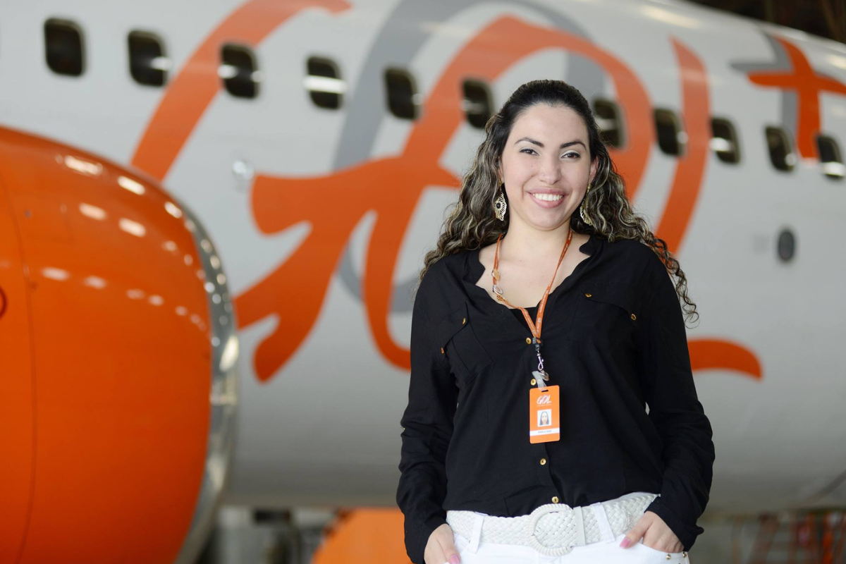 Emprego Gol Linhas Aéreas Mulher sorridente vestindo crachá da Gol frente a avião com logo laranja