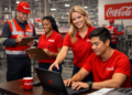Colaboradores da Coca-Cola usando uniformes vermelhos em ambiente industrial com pranchetas e laptop