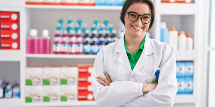 Mulher jovem sorridente com jaleco branco e óculos em farmácia com prateleiras cheias de medicamentos ao fundo