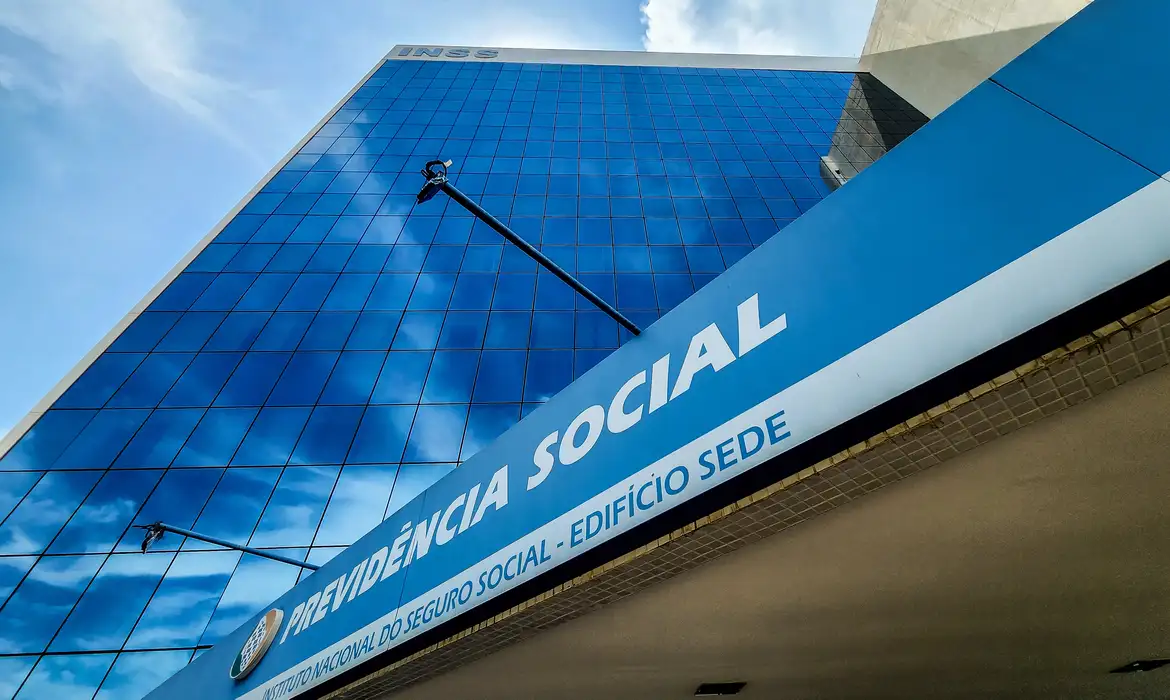 INSS e a antecipação do 13º salário de aposentados e pensionistas. Fachada do edifício sede do INSS com a inscrição 'Previdência Social' e reflexos do céu nas janelas, relacionado à antecipação do 13º salário para aposentados e pensionistas.