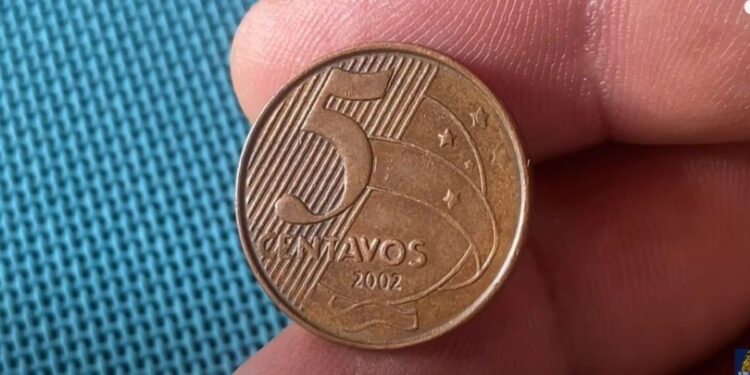 Close detalhado de uma moeda de 5 centavos de real do ano 2002, exibindo o número 5 e a data, segurada entre os dedos sobre um fundo azul texturizado.