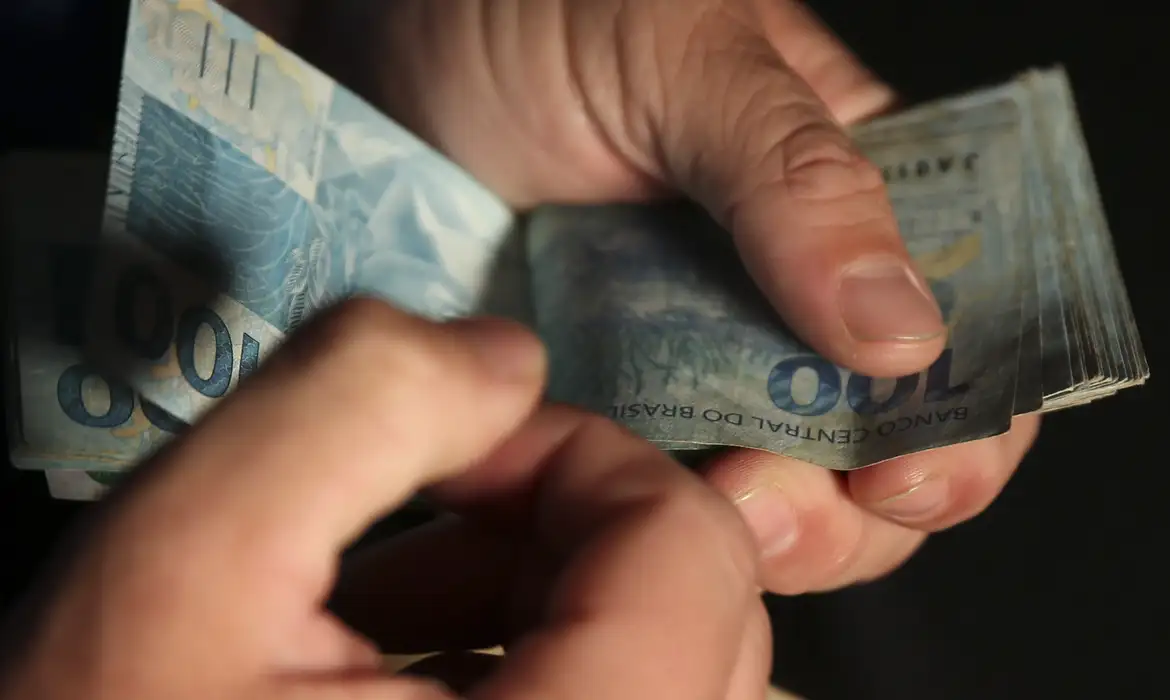 Bolsa Família 2026 Pessoa manuseando notas de R$ 100 do Banco Central do Brasil, representando o valor de R$ 950 que será pago neste sábado.