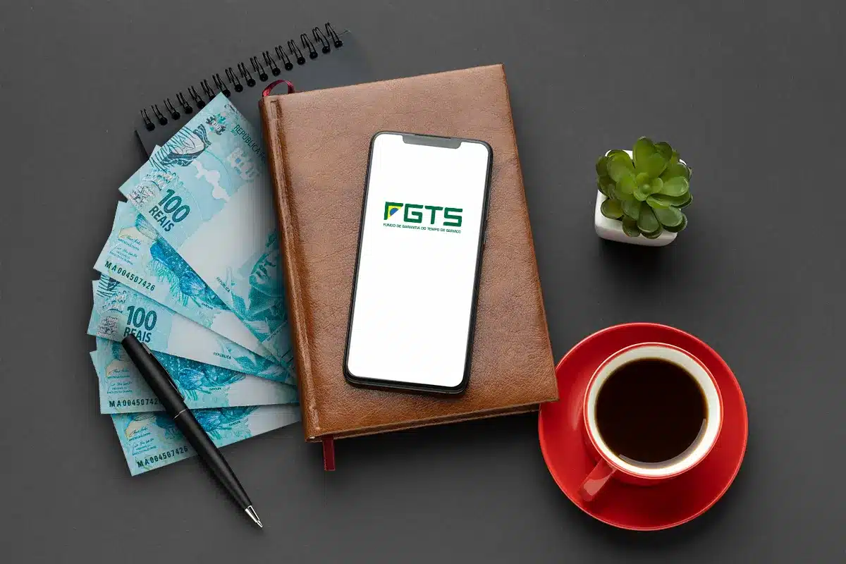 Calendário Saque aniversário do FGTS 2026 Celular com aplicativo FGTS sobre agenda e notas de 100 reais representando calendário do saque-aniversário 2026