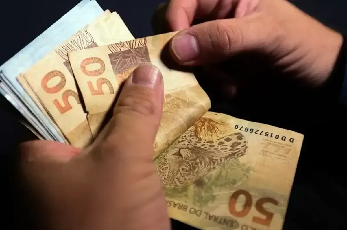 Calendário do INSS 2026 Mãos contando várias notas de cinquenta reais em fundo escuro
