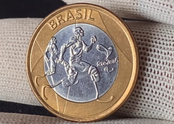 Close-up de uma mão segurando a moeda de 1 real das Paralimpíadas (Perna de Pau) entre os dedos, destacando o desenho do atleta.