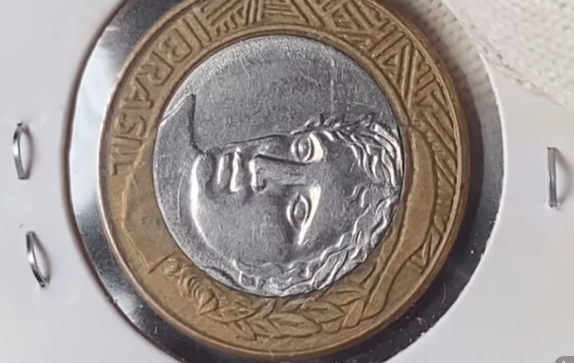 Close-up de uma moeda de 1 Real com a efígie da República centralizada, protegida por um suporte de acrílico branco com grampos laterais.