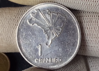 Close de uma moeda antiga de 1 Cruzeiro sendo segurada com cuidado por dedos usando luvas brancas, indicando seu potencial valor como item de colecionador raro.