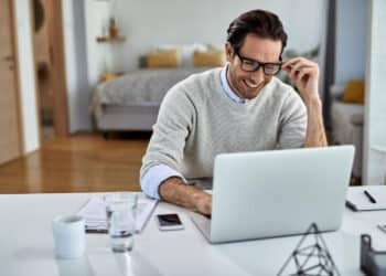 Profissional sorridente trabalhando em home office com notebook e celular em ambiente residencial confortável.