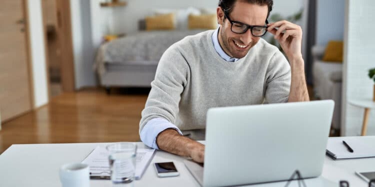 Profissional sorridente trabalhando em home office com notebook e celular em ambiente residencial confortável.