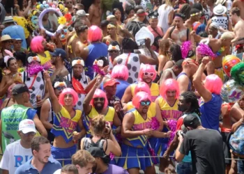 Bloco de Carnaval com foliões usando perucas cor-de-rosa e fantasias, destacando a data e as folgas de quem tem direito ao feriado.