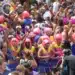Bloco de Carnaval com foliões usando perucas cor-de-rosa e fantasias, destacando a data e as folgas de quem tem direito ao feriado.