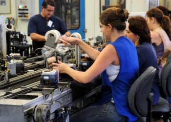 Trabalhadores em linha de produção industrial, realizando tarefas de montagem em ambiente de fábrica.