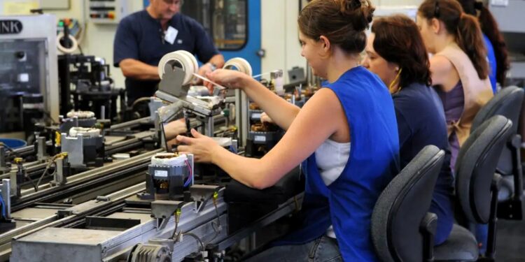 Trabalhadores em linha de produção industrial, realizando tarefas de montagem em ambiente de fábrica.