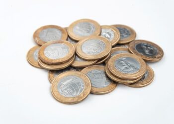 Pilha de moedas de 1 real de faces variadas, incluindo a efígie da República e a Cruz do Sul, sobre uma superfície branca limpa.