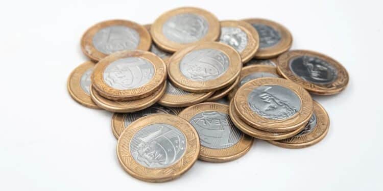 Pilha de moedas de 1 real de faces variadas, incluindo a efígie da República e a Cruz do Sul, sobre uma superfície branca limpa.