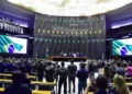 Sessão na Câmara dos Deputados com policial em destaque, em ambiente formal e institucional.