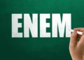 Mão segurando giz branco escrevendo a palavra "ENEM" em um quadro verde de escola.